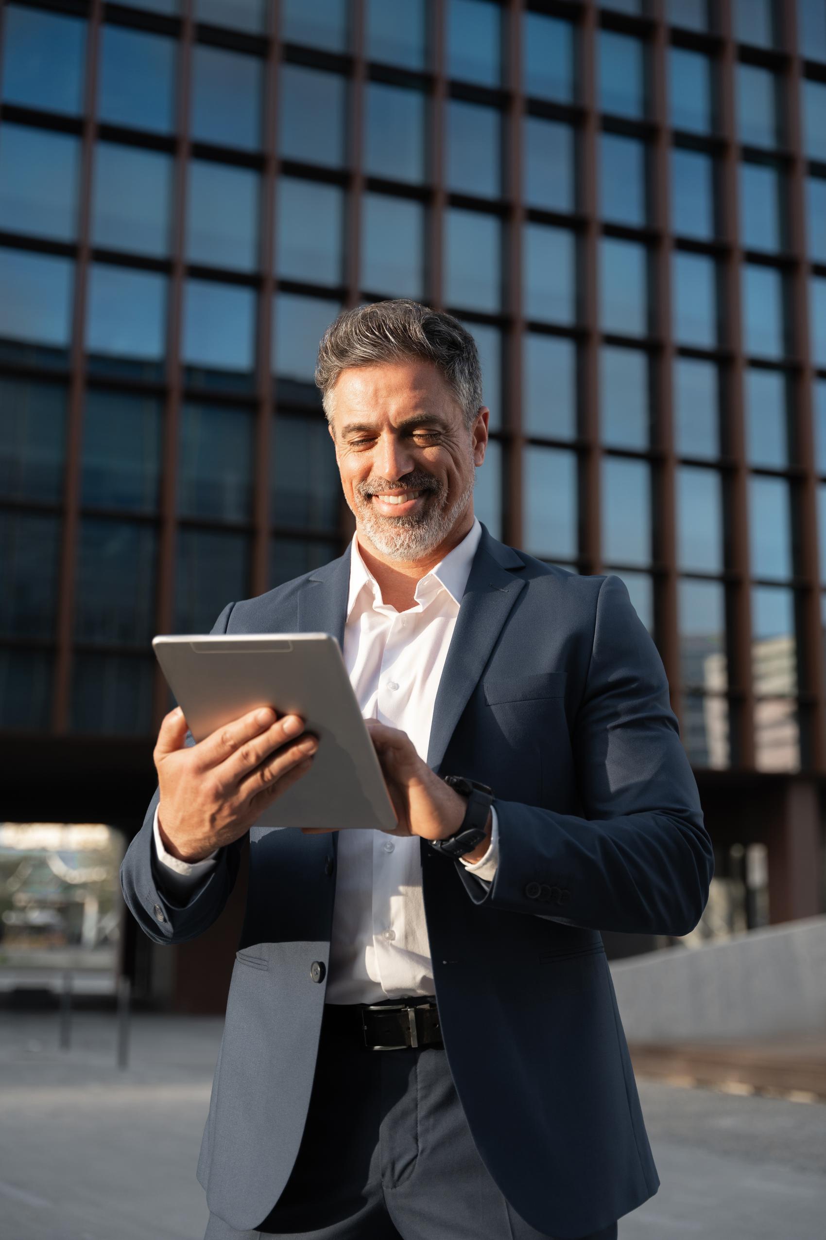 Zakelijke man in pak kijkt lachend naar een tablet voor een modern kantoorgebouw, passend bij financiële zekerheid mkb.