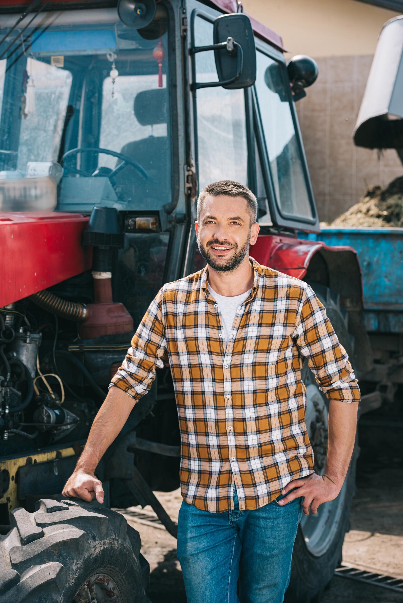 Boer in geruit overhemd staat met een glimlach naast zijn rode tractor, passend bij financieel advies voor mkb'ers.