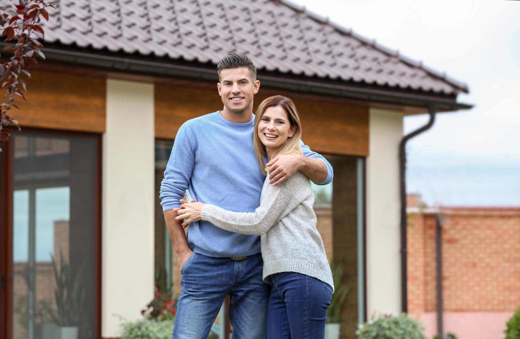 Jong stel staat glimlachend voor hun nieuwe woning, tevreden over hun hypotheek en actuele renteoverzicht.