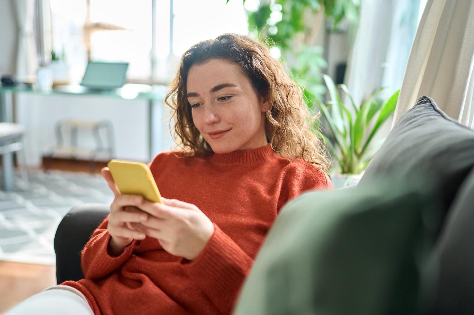 Vrouw in rode trui zit ontspannen op de bank en kijkt naar haar smartphone, en geeft een wijziging over haar polis door.